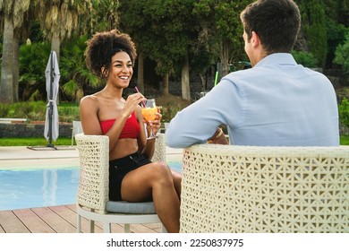 An African American woman sits by the pool, smiling and holding a glass of fruit smoothie while chatting with a young man. She is sitting on a straw chair - carefree and relaxing summer lifestyle. - Powered by Shutterstock