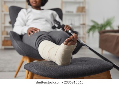 African american woman is sitting at home in armchair with broken bandaged leg lying on stool and holding crutches in her hands. Recovery time after physical injury of the foot bone. Selective focus.