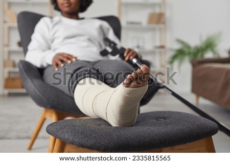 African american woman is sitting at home in armchair with broken bandaged leg lying on stool and holding crutches in her hands. Recovery time after physical injury of the foot bone. Selective focus.