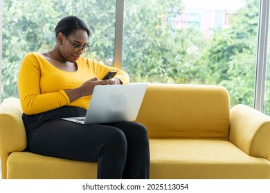 African American woman sitting on yellow sofa working on smartphone typing messages sending emails and working on laptop in living room at home concept quarantine and work from home 