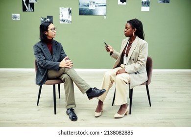 African American woman sitting on chair holding voice recorder interviewing modern young Asian photographer on first exhibition day in art gallery