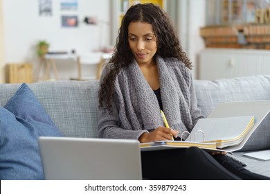 African American Woman Sitting On Her Sofa At Home Looking At Her Laptop And Taking Notes