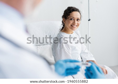 african american woman smiling near doctor in latex gloves holding thermometer on blurred foreground