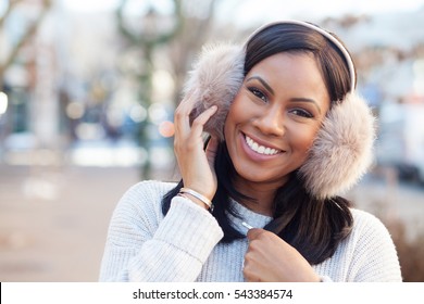 African American Woman Smiling Wearing Ear Muffs Downtown In The Winter With Head Tilted