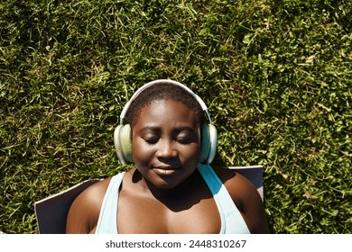 An African American woman in sportswear relaxes in nature, listening to music through headphones while laying in the grass.