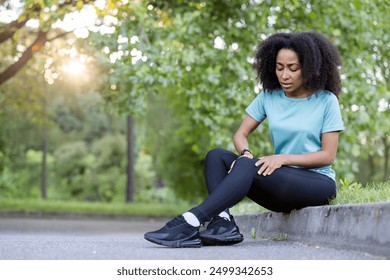 African American woman in sportswear sitting on roadside in park holding knee with a phone in hand. Concept of injury, exercise, and pain during workout. - Powered by Shutterstock