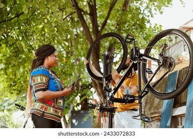 African american woman standing with digital tablet while caucasian man repairs damaged bike in yard. Two active persons outside using phone tablet for mending bicycle. - Powered by Shutterstock