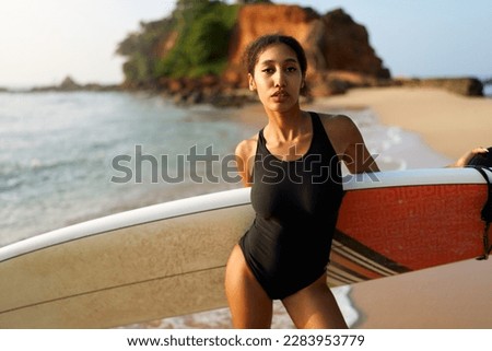 African american woman standing with surfboard on ocean beach. Black female surfer posing with surf board. Pretty multiethnic girl goes on surfing session on tropical location at sunny sunrise.