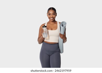 African American woman standing while holding a yoga mat in one hand and a water bottle in the other. She appears ready for a workout session or a yoga class, with a focused expression. - Powered by Shutterstock