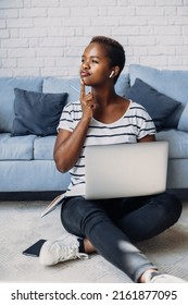 African American Woman Studying Remotely At Home Using A Computer Laptop Looking Away Thinking About Question, Sitting On The Floor. Curly Black Hair. Pensive