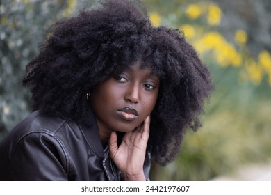 An African American woman with a stylish afro hairstyle gazes away thoughtfully. She is outdoors, with a backdrop of autumn leaves, providing a natural contrast that complements her look. Her hand - Powered by Shutterstock