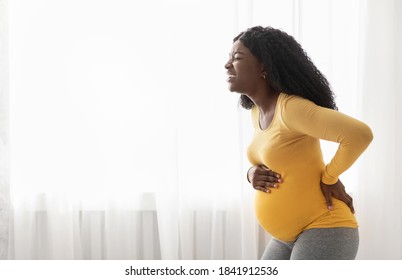 African american woman suffering from labor pains at home, standing by window, touching her tummy and back. Black expecting lady having childbirth labor, staying alone at home, copy space