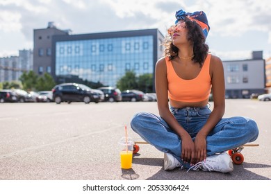 african american woman in sunglasses sitting with crossed legs on longboard near plastic cup with orange juice on asphalt