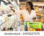 African American Woman Taking Milk Bottle From Shelf Holding Smartphone, Shopping Groceries In Supermarket On Weekend. Happy Female Customer Choosing Food Products In Grocery Shop