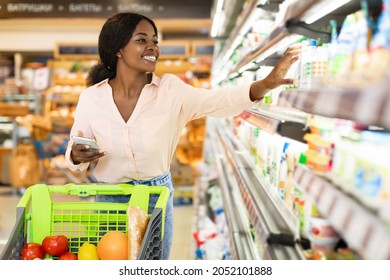 African American Woman Taking Milk Bottle From Shelf Holding Smartphone, Shopping Groceries In Supermarket On Weekend. Happy Female Customer Choosing Food Products In Grocery Shop
