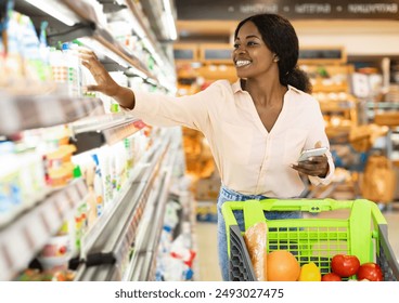 African American Woman Taking Milk Bottle From Shelf Holding Smartphone, Shopping Groceries In Supermarket On Weekend. Happy Female Customer Choosing Food Products In Grocery Shop - Powered by Shutterstock