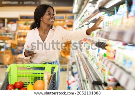 African American Woman Taking Milk Bottle From Shelf Holding Smartphone, Shopping Groceries In Supermarket On Weekend. Happy Female Customer Choosing Food Products In Grocery Shop