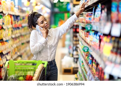 African American Woman Talking On Mobile Phone Buying Food During Grocery Shopping In Supermarket Store. Black Female Customer Choosing Groceries Chatting On Cellphone In Shop.