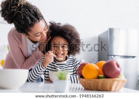 african american woman touching hair of happy kid eating corn flakes near fruits on blurred foreground