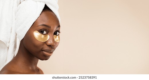 African American woman with a towel on her head and gold eye patches. The African American woman with towel on head and eye patches. Skincare routine for an African American woman. Beauty and skincare - Powered by Shutterstock