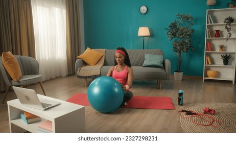 An African American woman in a tracksuit and headband sits crosslegged on a sports mat in the living room. A young woman holds a fitness ball in front of her and looks at a laptop on the table.