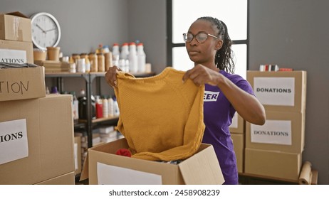 African american woman volunteering in a storage room, organizing yellow sweater in donation box. - Powered by Shutterstock