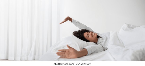 African American woman wakes up and stretches her arms wide while lying on a comfortable bed. Sunlight filters through the curtains, creating a warm atmosphere in her serene bedroom. - Powered by Shutterstock