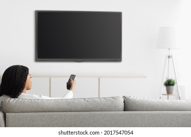 African American Woman Watching TV Pointing Remote Control At Flatscreen Plasma Television Set With Blank Screen, Switching Channels While Sitting On Sofa At Home, Mockup