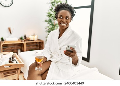 African american woman wearing bathrobe drinking champagne using smartphone at beauty salon - Powered by Shutterstock