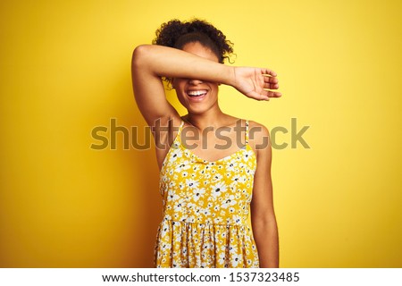 African american woman wearing casual floral dress standing over isolated yellow background covering eyes with arm smiling cheerful and funny. Blind concept.