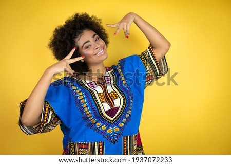 African american woman wearing african clothing over yellow background Doing peace symbol with fingers over face, smiling cheerful showing victory