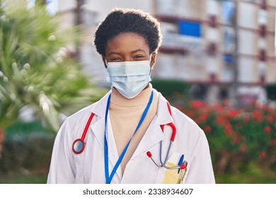 African american woman wearing doctor uniform and medical mask standing at park - Powered by Shutterstock