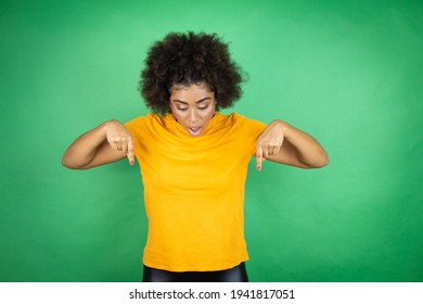 African american woman wearing orange casual shirt over green background surprised, looking down and pointing down with fingers and raised arms