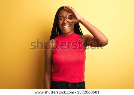 African american woman wearing red casual t-shirt standing over isolated yellow background doing ok gesture with hand smiling, eye looking through fingers with happy face.