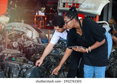 African American Woman And White Man Worker Working Together, Two People Checking Product Stock At Auto Spare Parts Store Shop Warehouse With Many Second Hand Engine Parts As Blurred Background