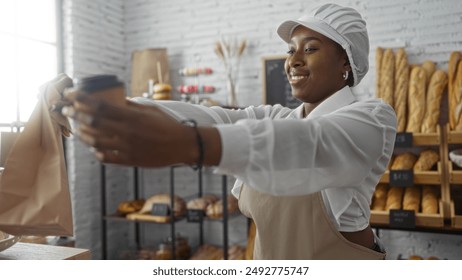 African american woman working in a bakery handing over a takeaway coffee and paper bag, smiling, in an indoor shop setting with bread and pastries in the background. - Powered by Shutterstock