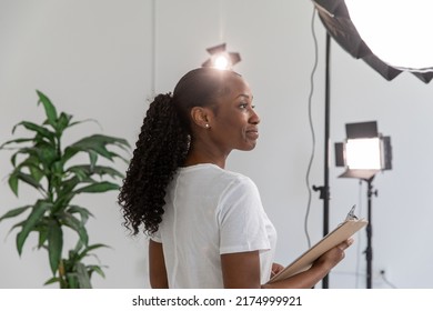 African American Woman Working On Film Set Holding Clipboard With Lighting And Equipment In Background. Proud Female Director In Video Production Studio