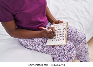 African American Woman Writing On Calendar Sitting On Bed At Bedroom