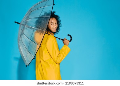 African American Woman In Yellow Waterproof Raincoat With Transparent Umbrella Isolated On Blue Studio Background. Autumn Season Concept