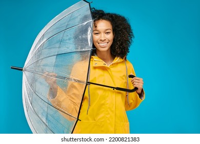 African American Woman In Yellow Waterproof Raincoat With Transparent Umbrella Isolated On Blue Studio Background. Autumn Season Concept