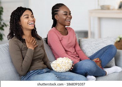 African American Women Watching Comedy At Home, Sitting On Sofa In Front Of TV And Eating Popcorn, Copy Space. Two Black Ladies Girlfriends Laughing, Enjoying Movie Together, Living Room Interior