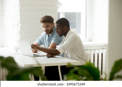African American worker explaining information and pointing at laptop to Caucasian colleague, both negotiating and discussing business strategy, working at computer in office. Teamwork, cooperation
