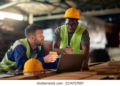 African American Worker And His Colleague Drinking Coffee While Working On Laptop At Wood Warehouse. 