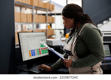 African american worker looking at inventory report on computer, working at merchandise quality control in storehouse. Stockroom supervisor preparing customers orders for shipping in warehouse
