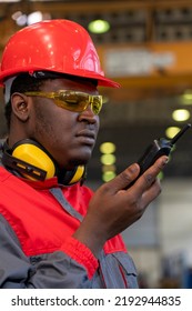 African American Worker In Protective Workwear Talking On Radio Communication Equipment In A Factory. Portrait Of Black Industrial Worker In Red Helmet, Safety Goggles And Safety Earmuffs.