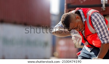 African American workers are exhausted from hard work. A container yard worker wearing a red reflective vest and hard hat is standing to wipe away sweat due to the hot weather.