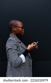 An African American young adult businesswoman is outside standing in front of a black wall and using her mobile phone. Sideview.