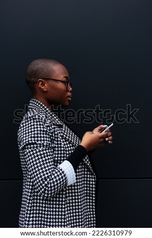 An African American young adult businesswoman is outside standing in front of a black wall and using her mobile phone. Sideview.