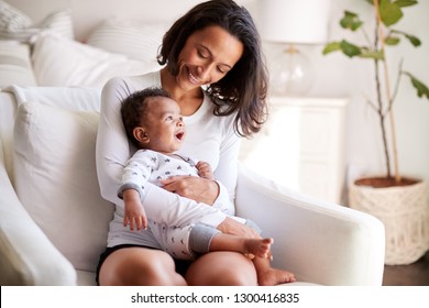African American young adult mother sitting in an armchair in her bedroom, holding her three month old baby son in her arms and looking down at him smiling, close up