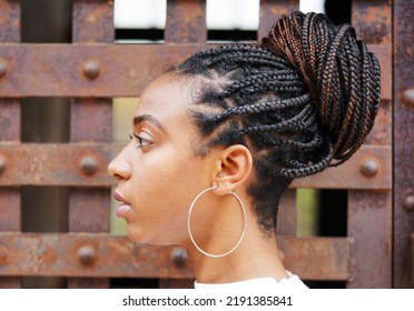 African American Young Attractive Woman Looks Left For A Profile Portrait Showing Off Her Braided Bun Hairstyle And Silver Hoop Earrings Against An Old Metal Door                               